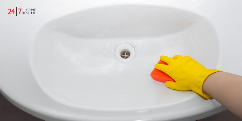 Hand in yellow glove cleaning a white sink with an orange sponge