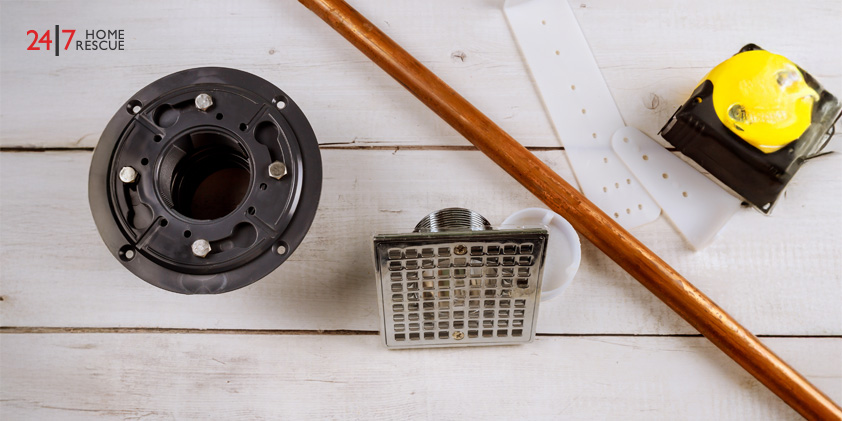 Plumbing tools including a black flange, metal drain cover, copper pipe, and yellow tape measure on a wooden surface