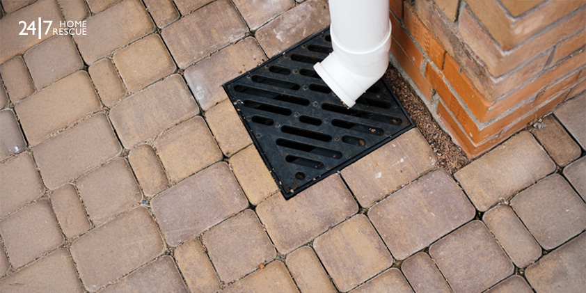 White downspout draining into a black grate on a brick-paved surface