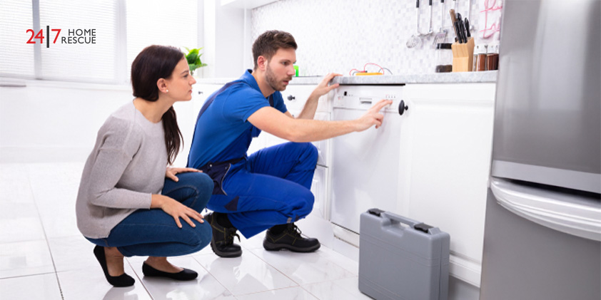 A serviceman crouching on the kitchen floor pressing the button of a dishwasher while a woman looks on
