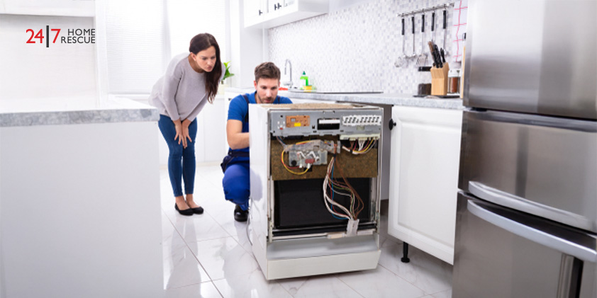 A smiling woman standing behind a technician repairing a dishwasher in the kitchen