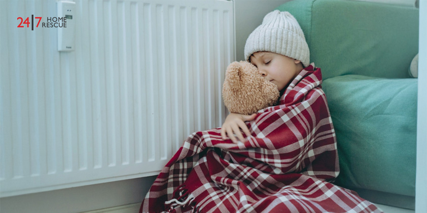 A child staying cosy by the radiator, wrapped in a blanket and holding a teddy bear for extra warmth
