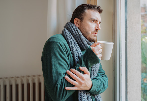 Man wrapped in a scarf holding a hot drink beside a cold radiator